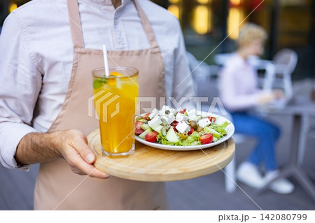 A server in an apron presents a fresh Greek salad and a refreshing orange beverage on a wooden tray at an outdoor cafe A server in an apron presents a fresh Greek salad and a refreshing orange beverage on a wooden tray at an outdoor cafe 142080799