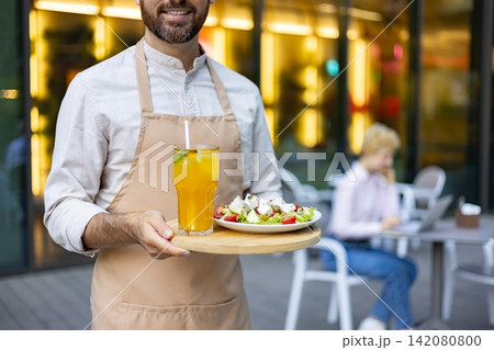 A smiling waiter in an apron holds a tray with a fresh salad and a refreshing beverage at an outdoor cafe A smiling waiter in an apron holds a tray with a fresh salad and a refreshing beverage at an outdoor cafe 142080800