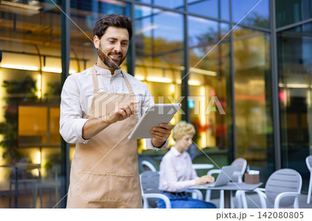 A smiling waiter in an apron uses a tablet to take an order outside a modern cafe with a glass facade A smiling waiter in an apron uses a tablet to take an order outside a modern cafe with a glass facade 142080805