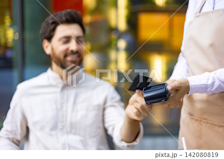 A man smiles while holding a credit card near a payment terminal held by a server wearing an apron A man smiles while holding a credit card near a payment terminal held by a server wearing an apron 142080819