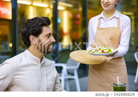 A smiling waiter presents a fresh salad to a happy customer at an outdoor cafe table with a refreshing drink A smiling waiter presents a fresh salad to a happy customer at an outdoor cafe table with a refreshing drink 142080829