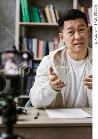 A man is sitting at a desk in front of a bookshelf, talking to a camera while recording a video A man is sitting at a desk in front of a bookshelf, talking to a camera while recording a video 142080955