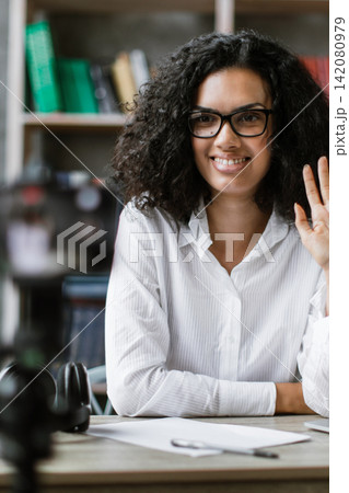 A young woman with curly hair and glasses smiles and waves hello while looking at the camera, positioned at her desk with a laptop and books 142080979