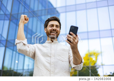A man with a beard smiles joyfully while holding his smartphone and raising his fist in a gesture of triumph outside a modern glass building A man with a beard smiles joyfully while holding his smartphone and raising his fist in a gesture of triumph outside a modern glass building 142081008