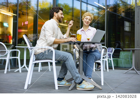 A man and woman are seated at a table outside a cafe, enjoying food and drinks while looking at a laptop 142081028