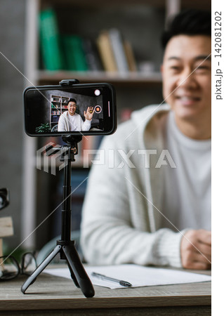 A man smiles and waves hello while recording a video on his smartphone, set up on a tripod in his home office A man smiles and waves hello while recording a video on his smartphone, set up on a tripod in his home office 142081202