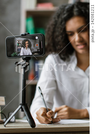 A young woman with curly hair is recording an online lesson using her smartphone on a tripod while writing notes at her desk A young woman with curly hair is recording an online lesson using her smartphone on a tripod while writing notes at her desk 142081211