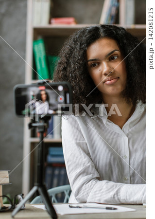A young woman with curly hair is actively engaged in a video call or online lesson, typing on her laptop while a smartphone records her A young woman with curly hair is actively engaged in a video call or online lesson, typing on her laptop while a smartphone records her 142081215