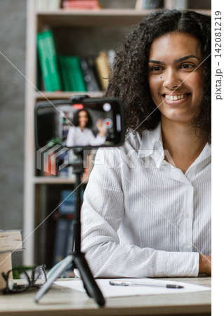 A young woman with curly hair smiles and waves while recording a video on her smartphone placed on a tripod in her home office 142081219