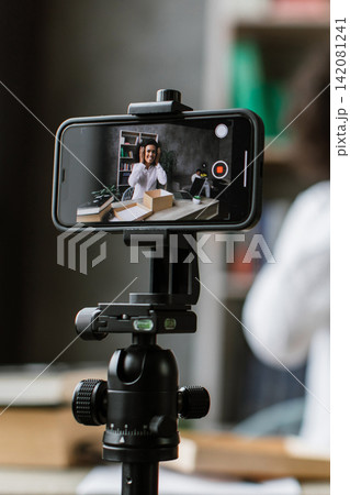 A woman records herself on a smartphone mounted on a tripod, preparing to share content online 142081241
