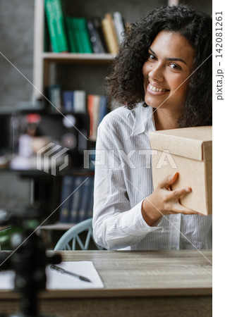 A smiling woman is recording herself unboxing a package for her online audience, showcasing a new product 142081261