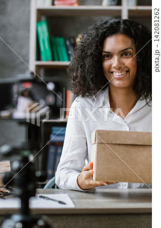 A young woman smiles while holding a cardboard box in front of a smartphone recording her content A young woman smiles while holding a cardboard box in front of a smartphone recording her content 142081262