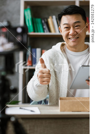 A smiling man gives a thumbs-up while holding a tablet and recording himself with a camera, suggesting content creation or online communication A smiling man gives a thumbs-up while holding a tablet and recording himself with a camera, suggesting content creation or online communication 142081279
