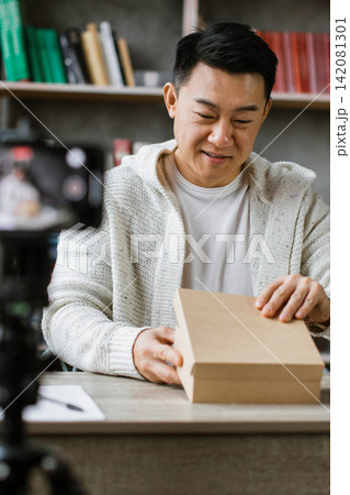 A man is recording himself opening a cardboard box while sitting at a desk with a laptop and books A man is recording himself opening a cardboard box while sitting at a desk with a laptop and books 142081301