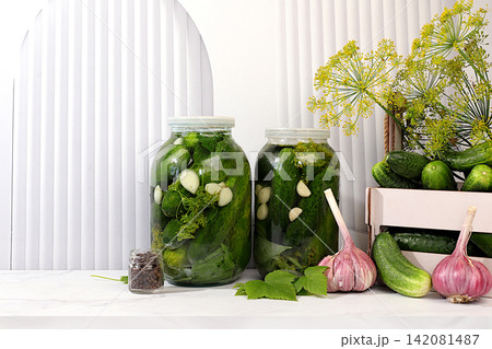 Cucumbers in a basket with dill, garlic, pickling leaves, and pickled cucumbers in jars on a kitchen counter. Harvest concept. Homemade pickle recipe and ingredients. Home canning concept. Cucumbers in a basket with dill, garlic, pickling leaves, and pickled cucumbers in jars on a kitchen counter. Harvest concept. Homemade pickle recipe and ingredients. Home canning concept. 142081487