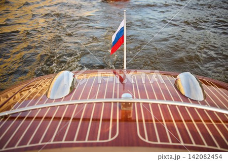 the glossy stern of a vintage pleasure boat at sunset, the boat in motion, bubbling water from under the boat, the flag of Russia fluttering in the wind, Neva river, varnished wood, chrome details the glossy stern of a vintage pleasure boat at sunset, the boat in motion, bubbling water from under the boat, the flag of Russia fluttering in the wind, Neva river, varnished wood, chrome details 142082454