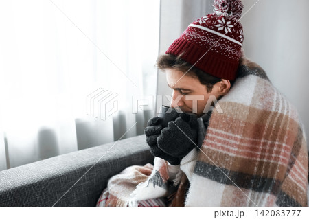 freezing man sits indoors wrapped in a blanket during a winter chill. Facing an energy crisis and high heating costs, he drinks a hot beverage to stay warm during the cold snap. freezing man sits indoors wrapped in a blanket during a winter chill. Facing an energy crisis and high heating costs, he drinks a hot beverage to stay warm during the cold snap. 142083777