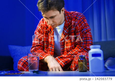 An unwell sick man sits at home in pajamas, suffering from a seasonal cold and flu infection. He looks at pills and medicine for respiratory illness treatment and recovery. 142083779