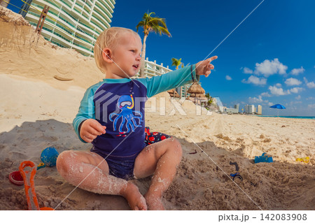 Three year old toddler playing with sand toys under sunny beach umbrella in Cancun, childhood, tropical family vacation and summer beach fun concept Three year old toddler playing with sand toys under sunny beach umbrella in Cancun, childhood, tropical family vacation and summer beach fun concept 142083908