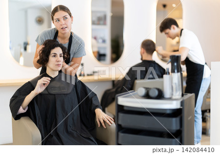 Young woman barbershop employee stands next to female client, discussing details of haircut Young woman barbershop employee stands next to female client, discussing details of haircut 142084410