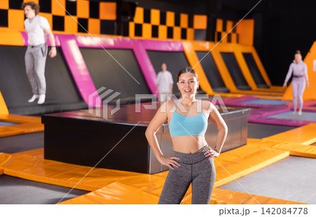 Happy young European female in blue top and gray leggings posing on trampolines during active weekend free time in sports center Happy young European female in blue top and gray leggings posing on trampolines during active weekend free time in sports center 142084778