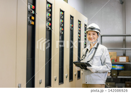 電子制御盤の前でタブレットを持つ作業服の女性 電子制御盤の前でタブレットを持つ作業服の女性 142086926