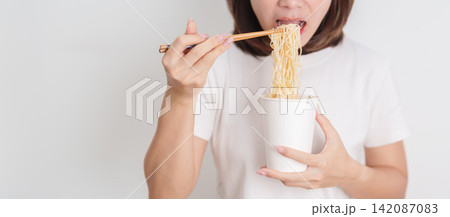 Happy young Asian woman enjoying a delicious cup of hot instant noodles while relaxing at home, Cheerful female eating instant ramen with a smile at apartment. Simple lifestyle and quick meal concept Happy young Asian woman enjoying a delicious cup of hot instant noodles while relaxing at home, Cheerful female eating instant ramen with a smile at apartment. Simple lifestyle and quick meal concept 142087083