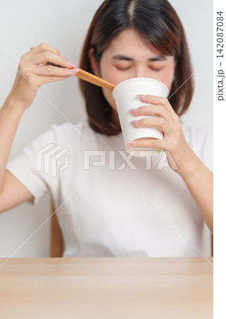 Happy young Asian woman enjoying a delicious cup of hot instant noodles while relaxing at home, Cheerful female eating instant ramen with a smile at apartment. Simple lifestyle and quick meal concept Happy young Asian woman enjoying a delicious cup of hot instant noodles while relaxing at home, Cheerful female eating instant ramen with a smile at apartment. Simple lifestyle and quick meal concept 142087084