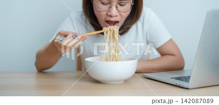 Happy young Asian woman working late at night on laptop while enjoying a bowl of hot instant ramen noodles at home. Cheerful female having a quick meal during work from home. Simple lifestyle concept Happy young Asian woman working late at night on laptop while enjoying a bowl of hot instant ramen noodles at home. Cheerful female having a quick meal during work from home. Simple lifestyle concept 142087089