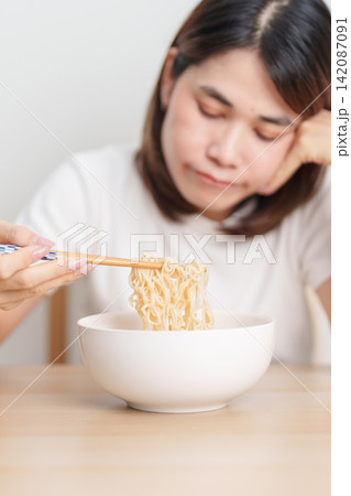 Bored young Asian woman eating instant noodles at home. Exhausted female looking unhappy while eating bowl ramen. quick meal after work, burnout and unhealthy eating habit, lifestyle and cheap meal Bored young Asian woman eating instant noodles at home. Exhausted female looking unhappy while eating bowl ramen. quick meal after work, burnout and unhealthy eating habit, lifestyle and cheap meal 142087091