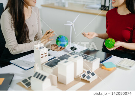 Two female architects discussing a green sustainable city model with solar panels and renewable energy concepts during a office meeting. Environmental 142090074