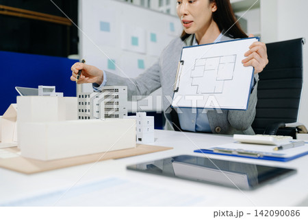 Female architect designing a sustainable urban building project model with solar panels, analyzing blue-print plans in office. 142090086
