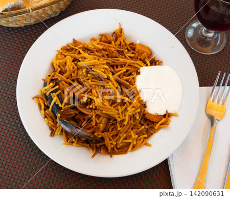 Fideua marinera(Spanish) seafood noodles with prawns and mussels served with sauce on a white plate in a restaurant 142090631