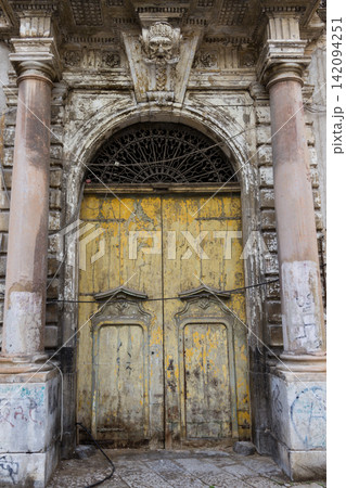 An ornate vintage yellow wooden entrance door framed by stone columns and an arched doorway featuring a carved grotesque mask keystone and intricate iron fanlight transom. An ornate vintage yellow wooden entrance door framed by stone columns and an arched doorway featuring a carved grotesque mask keystone and intricate iron fanlight transom. 142094251