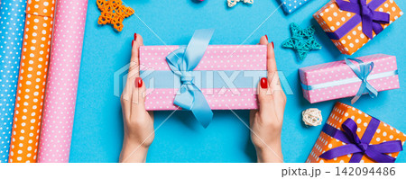 Top view of female hands holding a Christmas present on festive blue background. Holiday decorations and wrapping paper. New Year holiday concept Top view of female hands holding a Christmas present on festive blue background. Holiday decorations and wrapping paper. New Year holiday concept 142094486