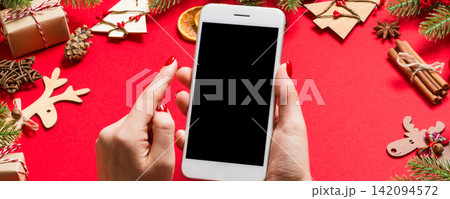 Top view of phone in female hand on festive red background. Christmas decorations. New Year time holiday. Mockup 142094572