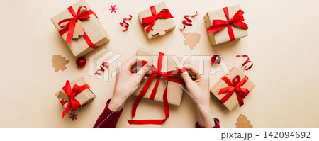 Woman is packing surprise gift for Christmas holidays, sitting at workplace with craft tools, top view. Woman wrapping Christmas gift at colored table Woman is packing surprise gift for Christmas holidays, sitting at workplace with craft tools, top view. Woman wrapping Christmas gift at colored table 142094692