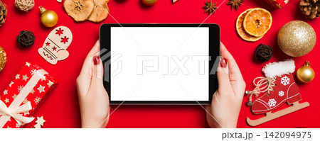 Top view of woman holding tablet in her hands on red background made of Christmas decorations. New Year holiday concept. Mockup 142094975