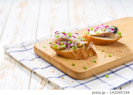Slices of baguette with pieces marinated herring fillet with with red onions and pickles on table. Slices of baguette with pieces marinated herring fillet with with red onions and pickles on table. 142095194