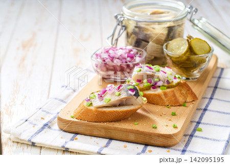 Slices of baguette with pieces marinated herring fillet with with red onions and pickles on table. Slices of baguette with pieces marinated herring fillet with with red onions and pickles on table. 142095195