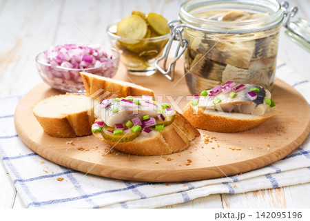 Slices of baguette with pieces marinated herring fillet with with red onions and pickles on table. Slices of baguette with pieces marinated herring fillet with with red onions and pickles on table. 142095196
