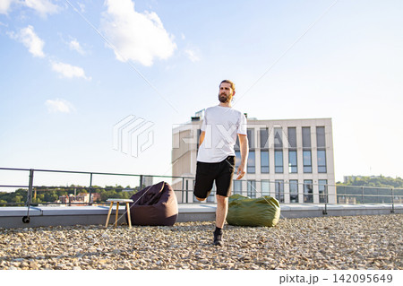 A man with a beard and tied-back hair performs a quadriceps stretch on a gravel rooftop with a modern building in the background 142095649