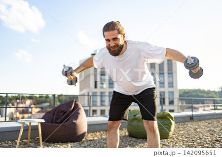 A man with a beard and bun hairstyle is performing a dumbbell workout on a rooftop terrace with a city view in the background A man with a beard and bun hairstyle is performing a dumbbell workout on a rooftop terrace with a city view in the background 142095651