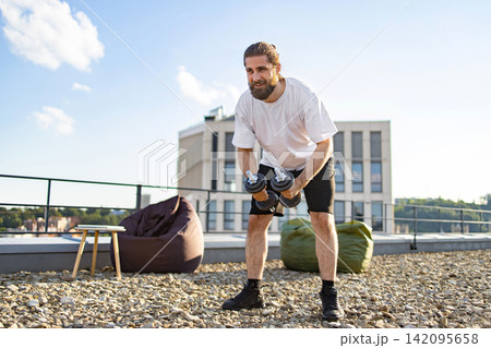 A fit man with a beard performs dumbbell exercises on a rooftop terrace with beanbags and a city view A fit man with a beard performs dumbbell exercises on a rooftop terrace with beanbags and a city view 142095658