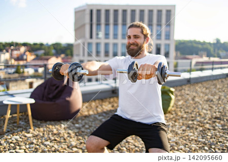 A smiling man with a beard and long hair is performing bicep curls with dumbbells on a rooftop terrace with a city view 142095660