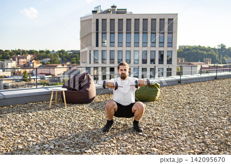 A bearded man with long hair performs dumbbell squats on a rooftop terrace with a city view and beanbag chairs 142095670