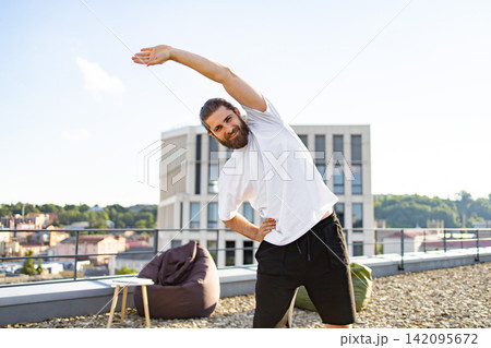 A man with a beard and ponytail smiles while stretching his body on a rooftop with a city view and modern building in the background A man with a beard and ponytail smiles while stretching his body on a rooftop with a city view and modern building in the background 142095672