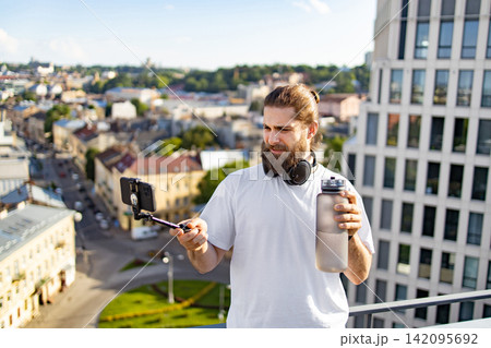 A bearded man with headphones around his neck records himself on a smartphone attached to a selfie stick while holding a large water bottle on a rooftop overlooking a city 142095692