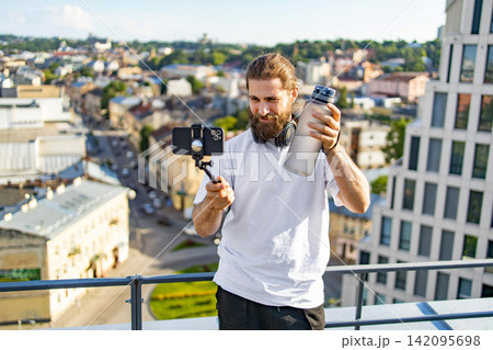 A man with a beard and headphones films himself with a smartphone on a selfie stick while holding a water bottle on a rooftop overlooking a city A man with a beard and headphones films himself with a smartphone on a selfie stick while holding a water bottle on a rooftop overlooking a city 142095698