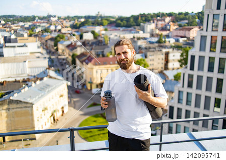 A bearded man in a white t-shirt holds a yoga mat and water bottle while standing on a rooftop overlooking a city A bearded man in a white t-shirt holds a yoga mat and water bottle while standing on a rooftop overlooking a city 142095741